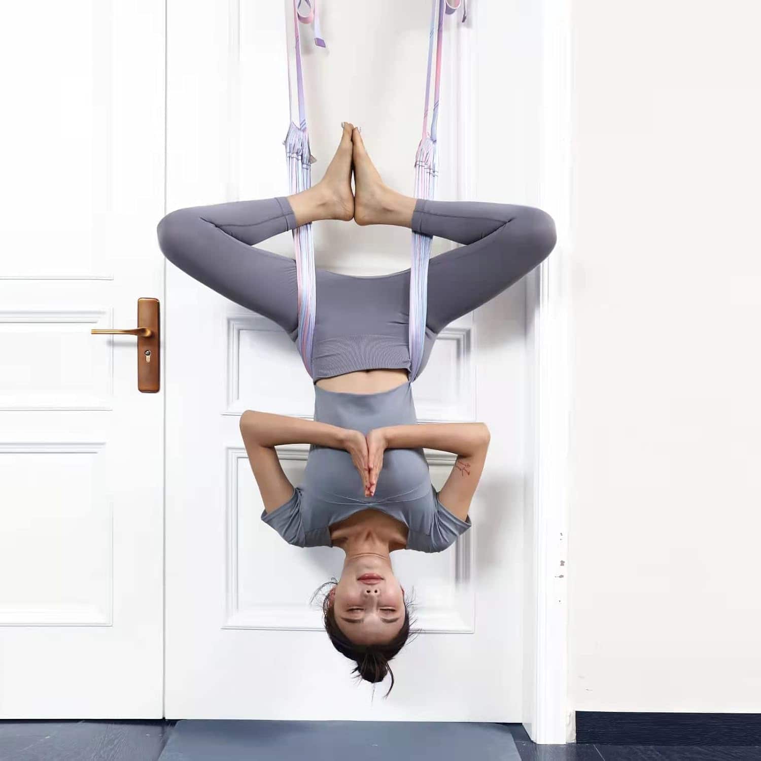 Person in gray athletic wear using an aerial yoga hammock against a white door.